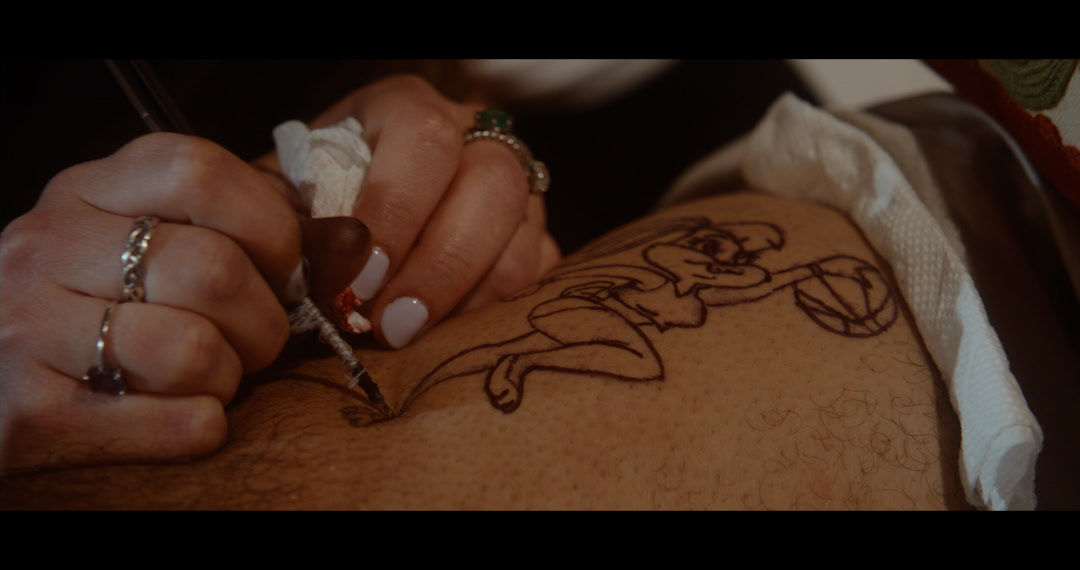 A close cropped shot showing a pair of hands decorated with rings and white nail polish are holding a syringe and injecting into what appears to be a thigh with a tattoo of Lola Bunny playing basketball